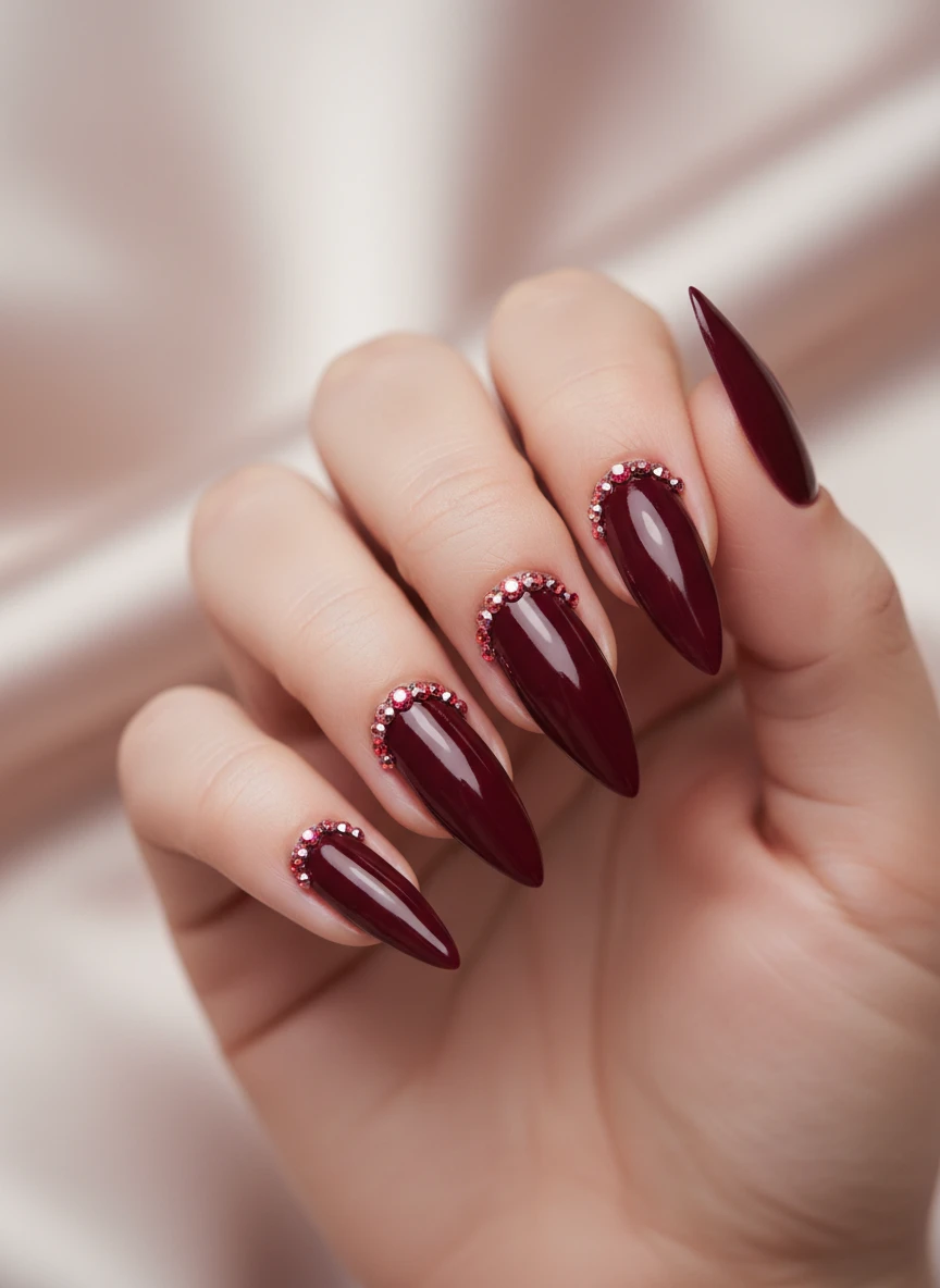 Elegant burgundy stiletto nails featuring a crown-like arrangement of tiny red and pink rhinestones.
