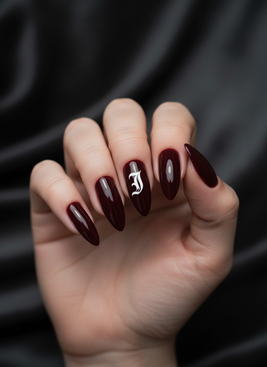 </strong> Close-up detail of long stiletto nails painted in deep burgundy wine color, featuring a bold Old English font initial 'J' in white on the middle finger, glossy top coat, moody dramatic lighting, fashion editorial style.